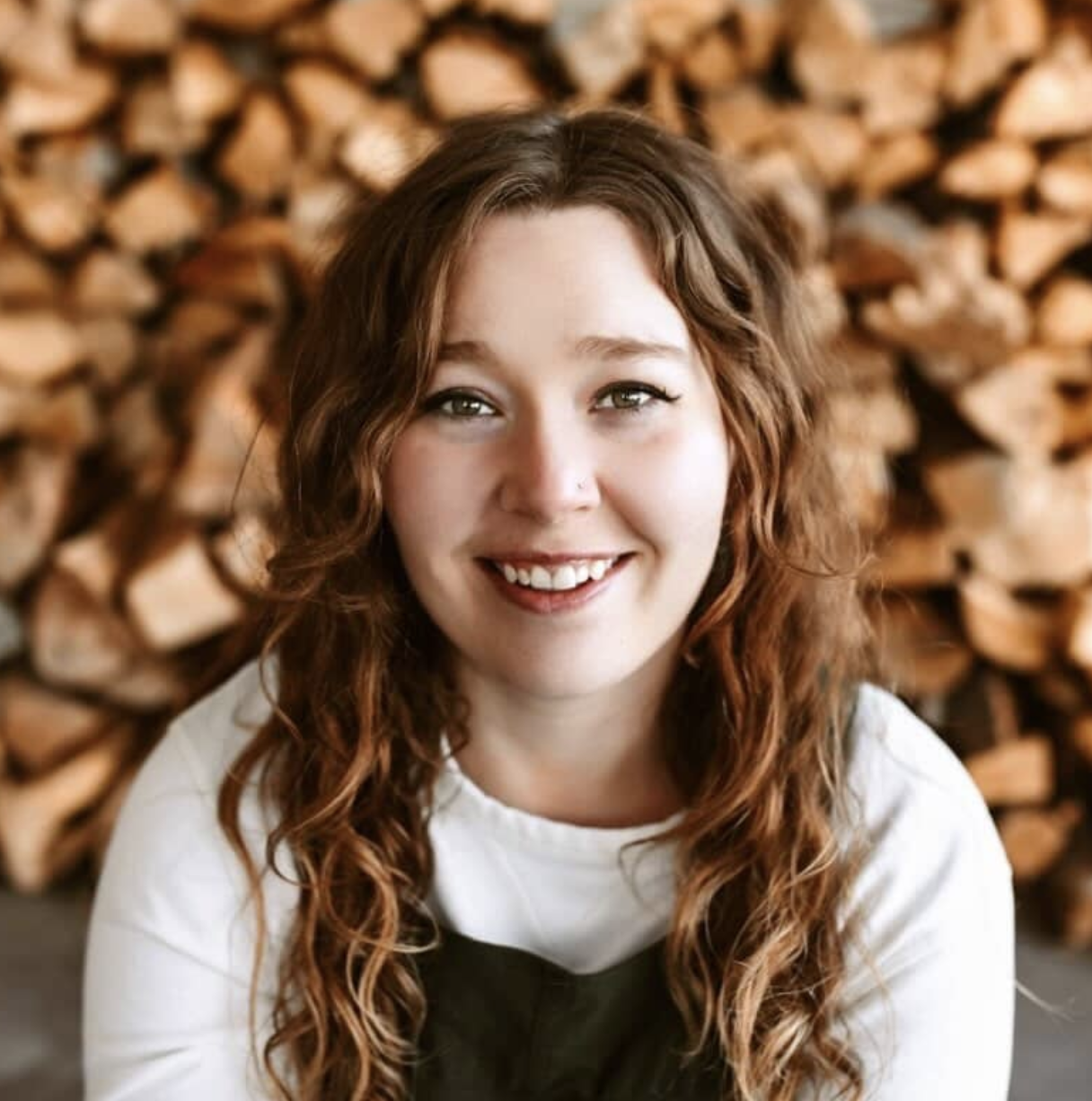 Image of a woman with hazel eyes, brown wavy hair, smiling softly, wearing a white long sleeve shirt and charcoal overalls